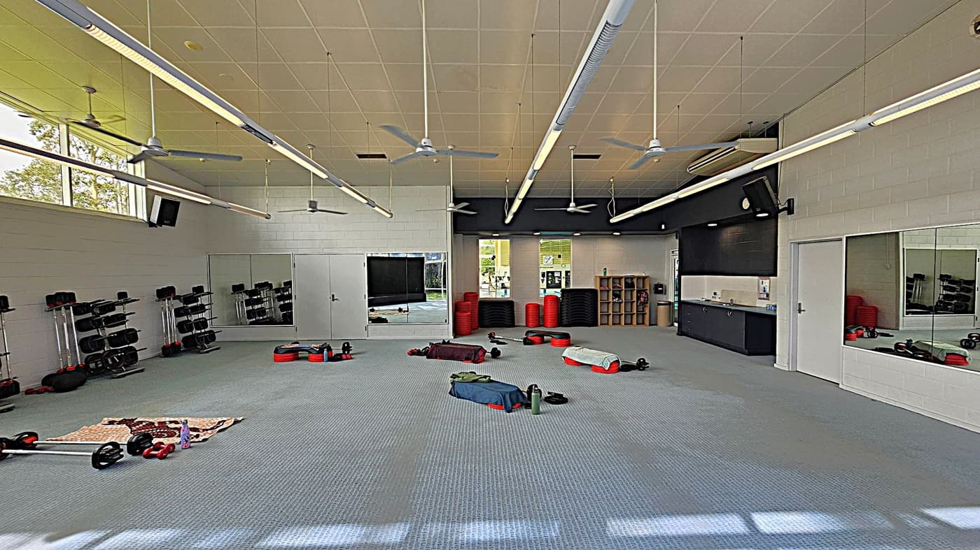 A spacious gym room with a grey carpeted floor. On the left, there are racks of weights and exercise equipment. In the centre, various exercise mats and weights are scattered. The right side features large mirrors and a black counter with storage shelves. The ceiling has multiple ceiling fans and long fluorescent lights. Windows in the background let in natural light, and there are red exercise steps stacked near the back wall.