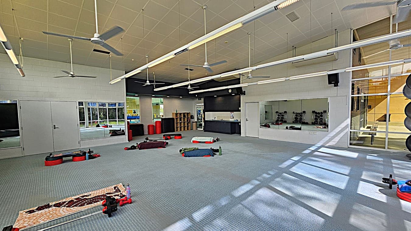 A spacious gym room with a light blue carpeted floor. On the left, there are large mirrors and a closed double door. In the centre, exercise equipment like weights and benches are scattered. The right side features a large window with sunlight streaming in, casting shadows on the floor. The ceiling has several ceiling fans and long fluorescent lights. In the background, there are shelves with red exercise mats and a small kitchenette area.