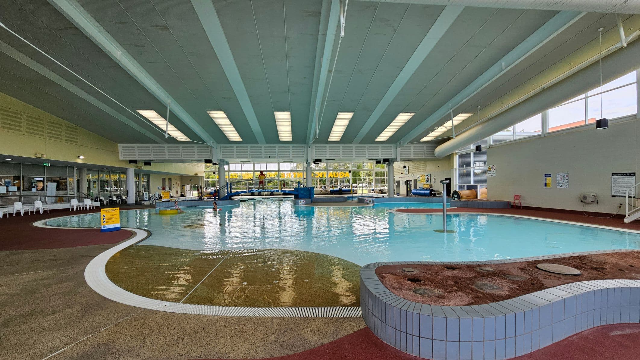 Indoor swimming pool with a curved design. On the left, a lifeguard stands on a blue platform near a small slide. Large windows with yellow signage allow natural light in. The right side features a water slide and a shallow area with a yellow sign. The ceiling has a series of skylights, and the floor is a mix of wet and dry areas with safety markings.