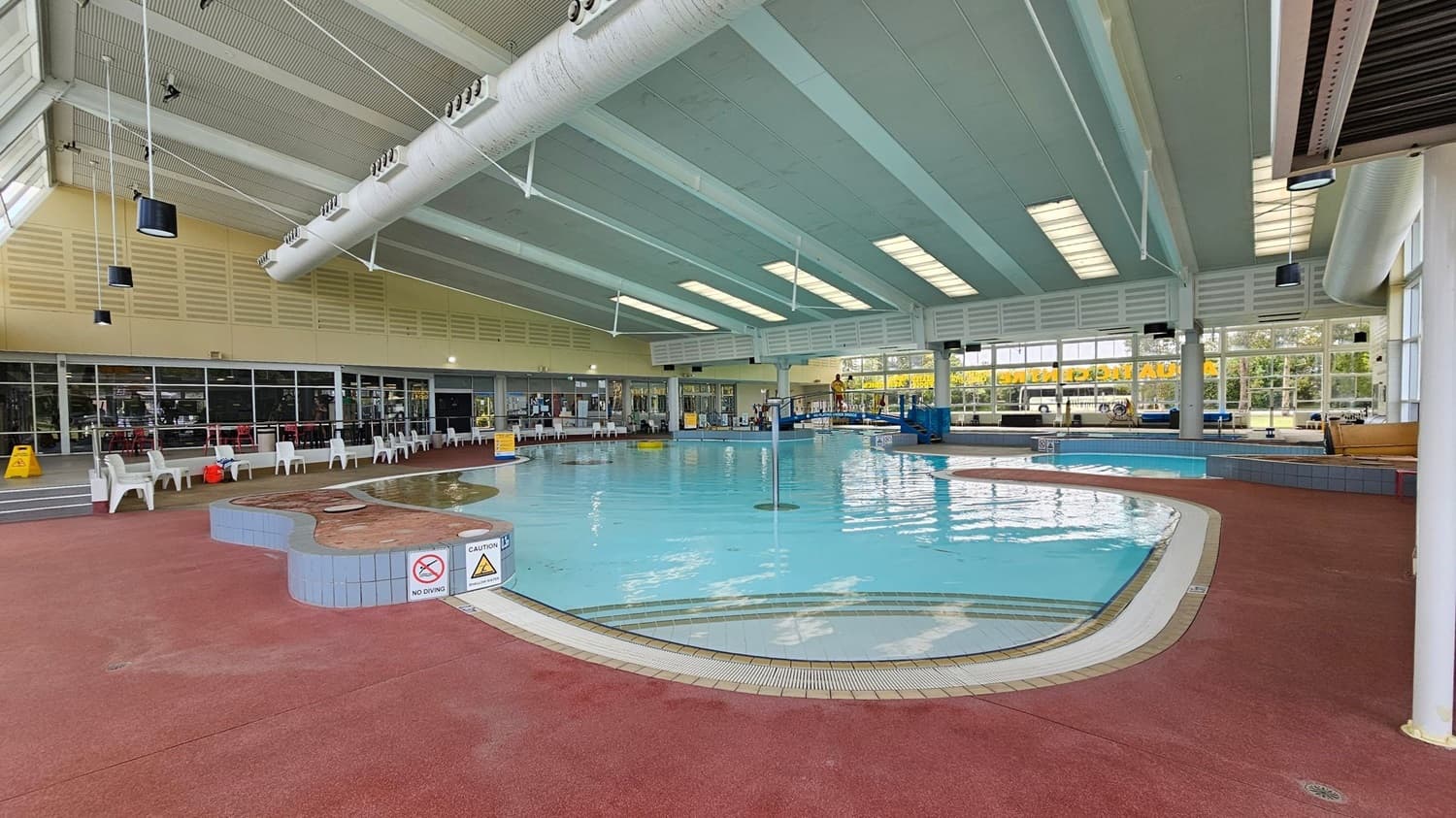 Indoor swimming pool with a light blue ceiling featuring skylights. The pool is surrounded by a red non-slip floor. On the left, there are white plastic chairs lined up against a wall with large windows. In the background, a lifeguard sits on a chair near the pool. Signs indicating "No Diving" and "Caution" are visible near the pool's edge. Large windows on the right let in natural light, and a bus is visible outside.