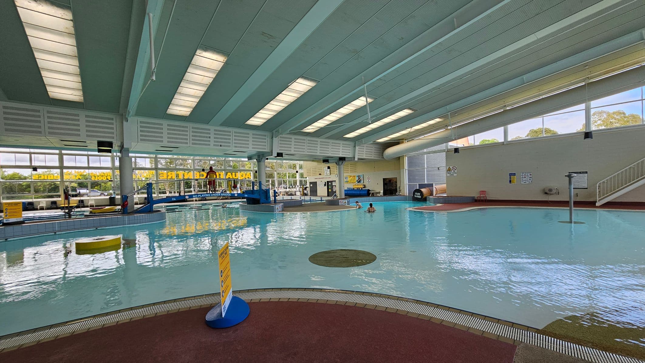 Indoor swimming pool with a shallow area in the foreground. The pool has a curved edge with a brown non-slip floor surrounding it. On the left, large windows let in natural light, and there are signs and a lifeguard chair. In the background, a few people are in the water near a small bridge. The ceiling is high with fluorescent lights, and a staircase is visible on the right leading to a slide. The walls are light-coloured with safety signs.