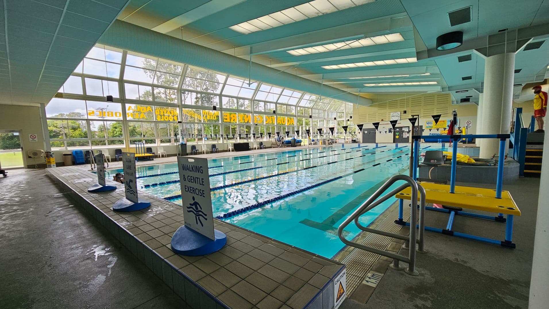 Indoor swimming pool with lanes marked by blue ropes. On the left, signs indicate "Walking & Gentle Exercise" and "Slow Lane." Large windows on the left wall provide natural light and a view of trees outside. The right side features a yellow and blue pool lift and a lifeguard standing near steps. The ceiling has rectangular lights, and the floor is wet with a non-slip surface.