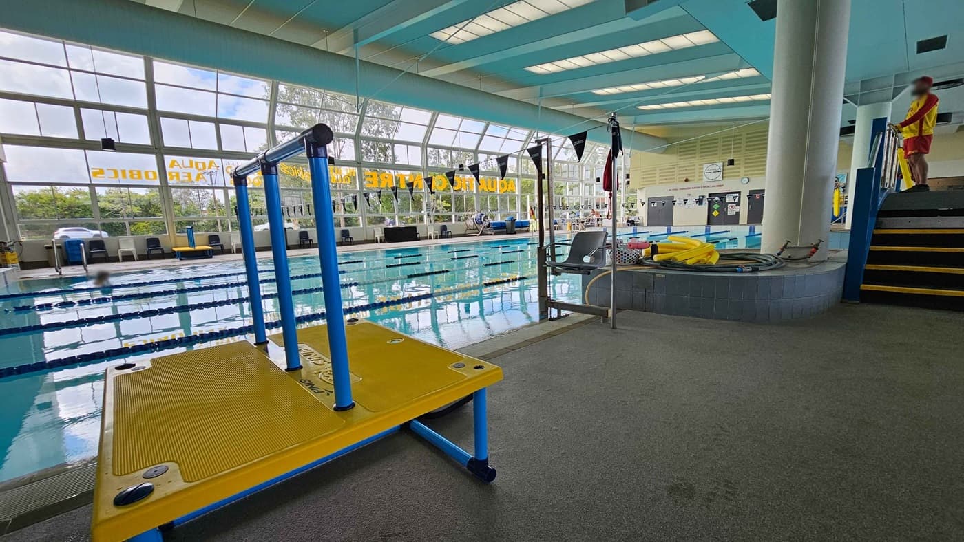 Indoor swimming pool with large windows on the left, allowing natural light in. A swimmer is in the pool, which has lane dividers. On the right, a lifeguard in red and yellow stands on a platform near a hot tub. A yellow and blue platform is in the foreground. The ceiling has skylights, and the floor is grey with some green patches. Various pool equipment is visible near the hot tub.