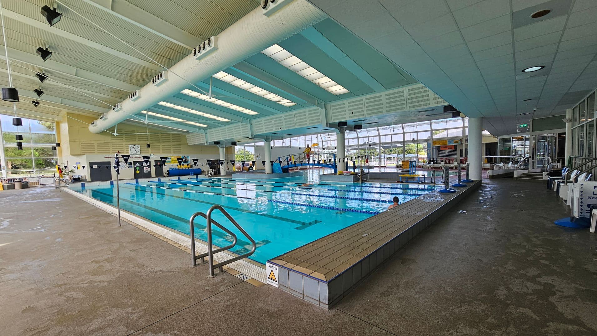 Indoor swimming pool with lanes, surrounded by a tiled deck. On the left, large windows let in natural light, and a clock is mounted on the wall. The ceiling is high with exposed beams and lighting fixtures. On the right, there are steps leading to a seating area with chairs. The pool is divided into lanes with a few swimmers. The floor is a mix of wet and dry areas, and there are caution signs near the pool's edge.