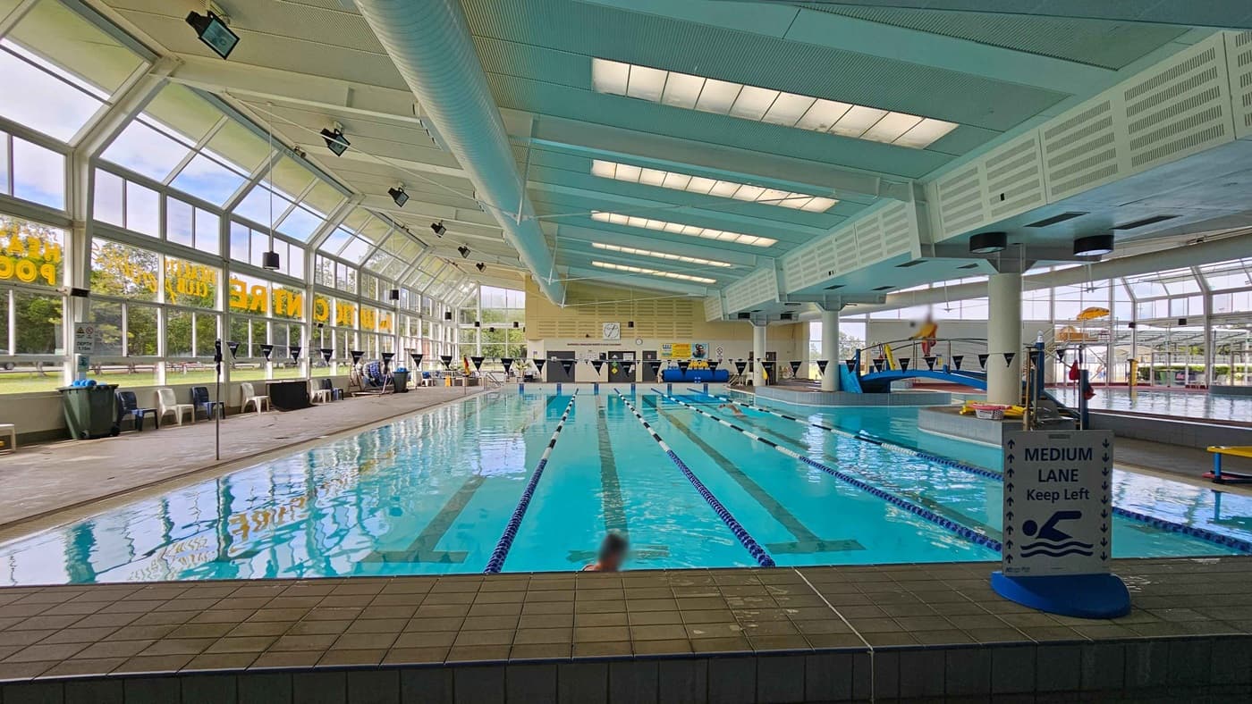 Indoor swimming pool with multiple lanes, marked by blue ropes. A sign in the foreground reads "Medium Lane Keep Left." On the left, large windows with yellow lettering allow natural light in, with chairs and bins along the wall. The ceiling is high with skylights and ventilation ducts. In the background, a lifeguard sits near a diving board, and there are additional pool areas visible. The floor is tiled around the pool's edge.