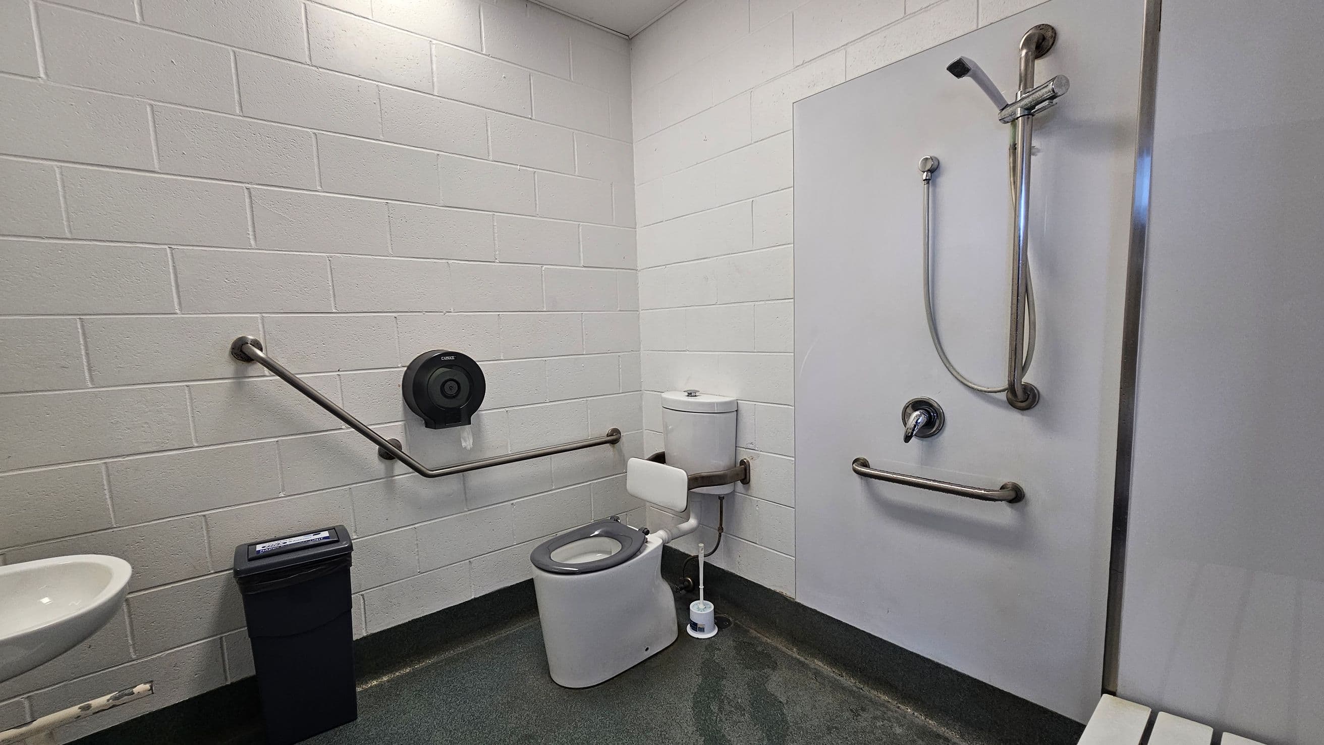 A bathroom with white brick walls and a grey floor. On the left, there's a white sink and a black bin. A metal grab bar runs along the wall, with a toilet paper dispenser above it. In the centre, a toilet with a white seat and a support bar is positioned against the wall. On the right, a shower area with a handheld showerhead and another grab bar is visible. A small brush is on the floor near the toilet. The ceiling is plain and white.