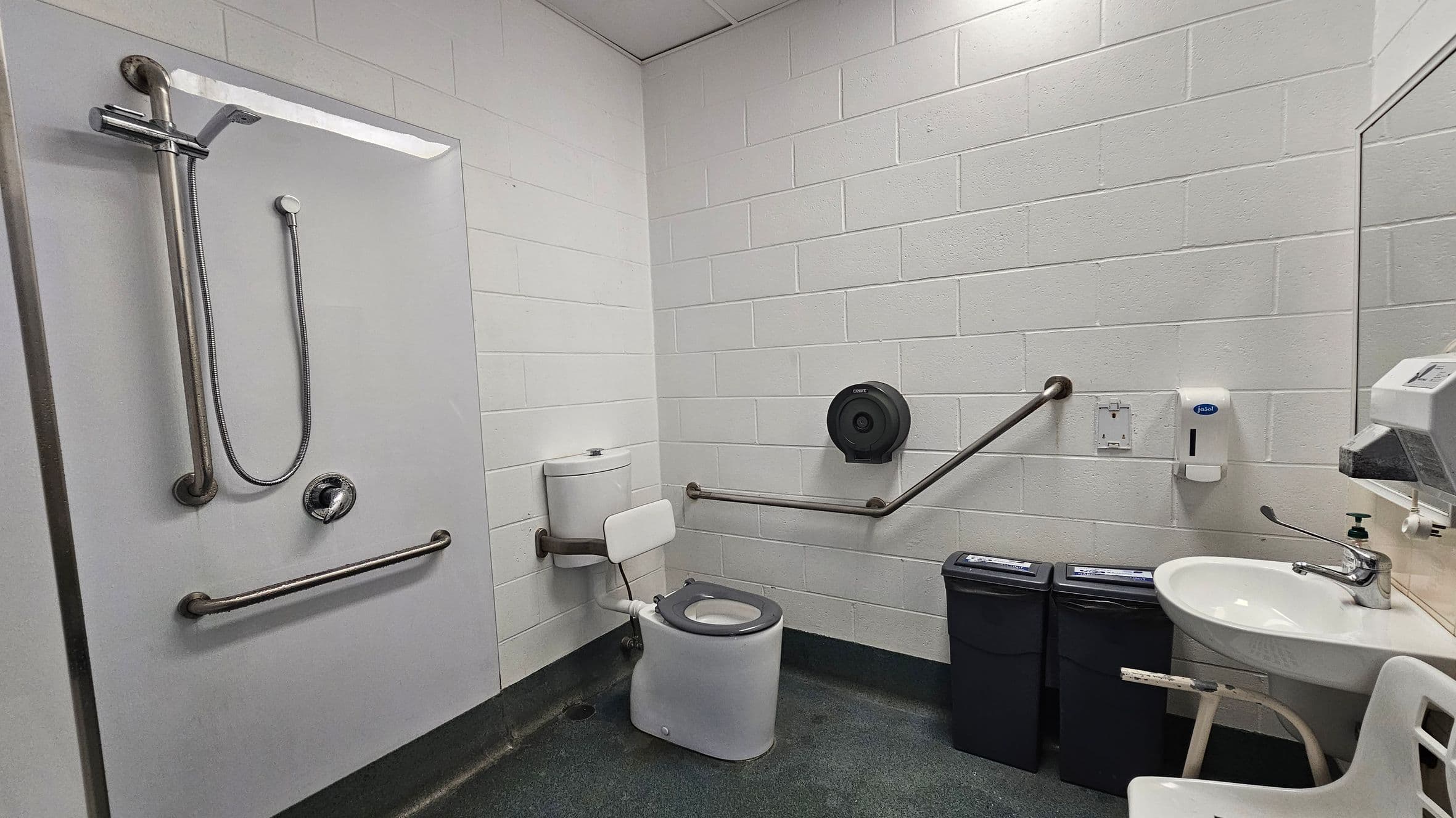 A public accessible bathroom with white brick walls and a grey floor. On the left, there's a shower area with a handheld showerhead and grab bars. In the centre, a toilet with a backrest and grab bars on both sides. A toilet paper dispenser is mounted on the wall. On the right, a sink with a lever tap, a soap dispenser, and a paper towel dispenser. Below the sink are two black bins. A white chair is partially visible in the foreground.