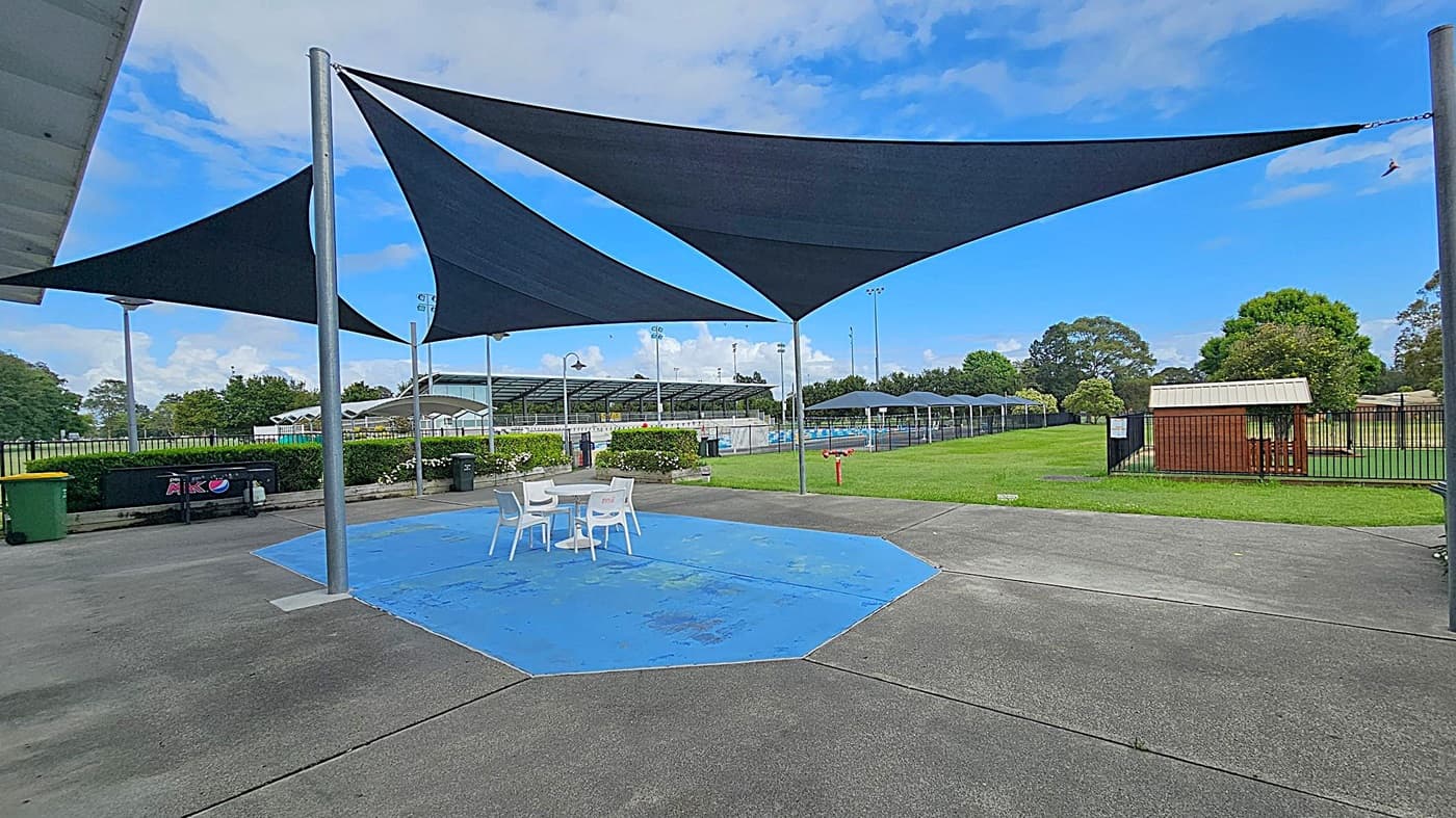 A concrete area with a blue-painted section in the centre, featuring a white table and chairs. Overhead, there are triangular shade sails supported by metal poles. To the right, a grassy area with a small wooden shed and trees. In the background, a fenced swimming pool with covered seating and more trees. A green bin and a vending machine are on the left side.