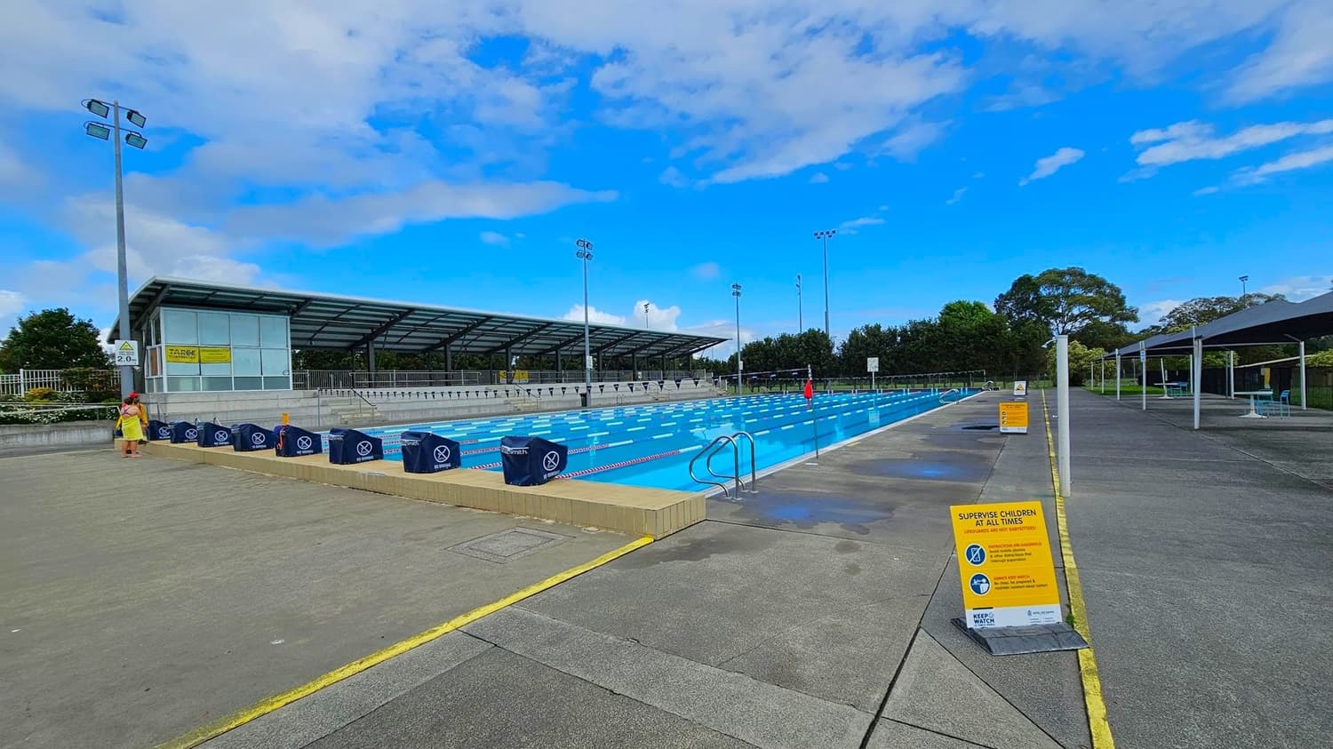 An outdoor swimming pool with multiple lanes is in the centre, surrounded by a concrete deck. On the left, there's a covered seating area with a glass-fronted building and a person standing nearby. On the right, there are signs with safety instructions and a few shaded seating areas. Tall floodlights are visible around the pool, and trees line the background. The pool has a metal ladder on the right side.
