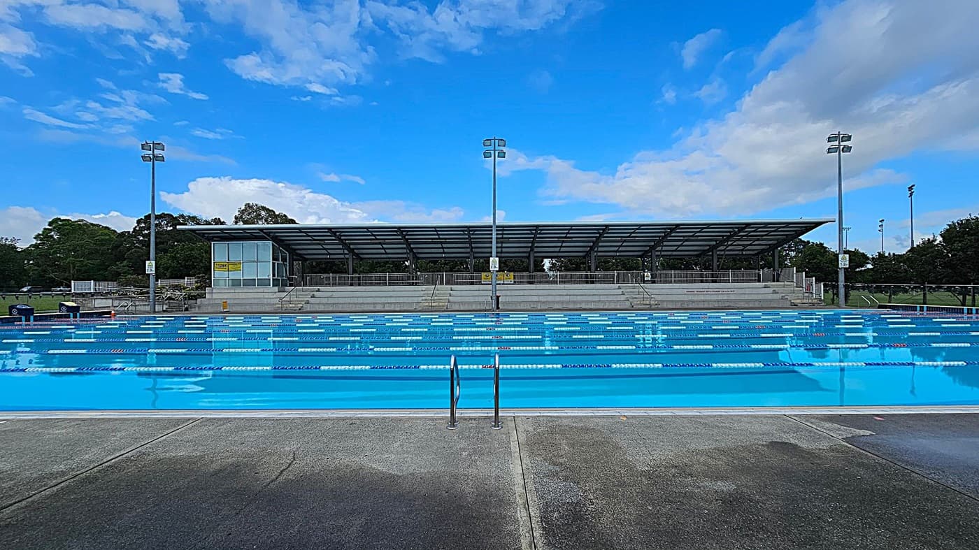 An outdoor swimming pool with clear blue water and lane dividers stretches across the foreground. In the background, there is a covered seating area with a metal roof and several rows of benches. Tall floodlights are positioned on either side of the pool. Trees and greenery are visible beyond the seating area. The pool deck is made of concrete, and there are metal handrails leading into the water.