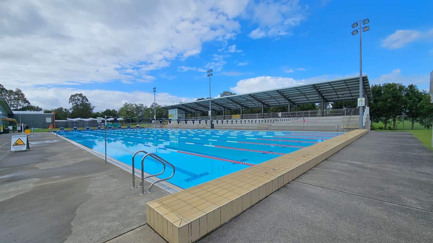 An outdoor swimming pool with multiple lanes, bordered by a tiled edge. On the left, a caution sign warns of a sudden drop. The pool is surrounded by a concrete deck. In the background, there is a covered seating area with a metal roof and several tall floodlights. Trees and greenery are visible beyond the seating area.