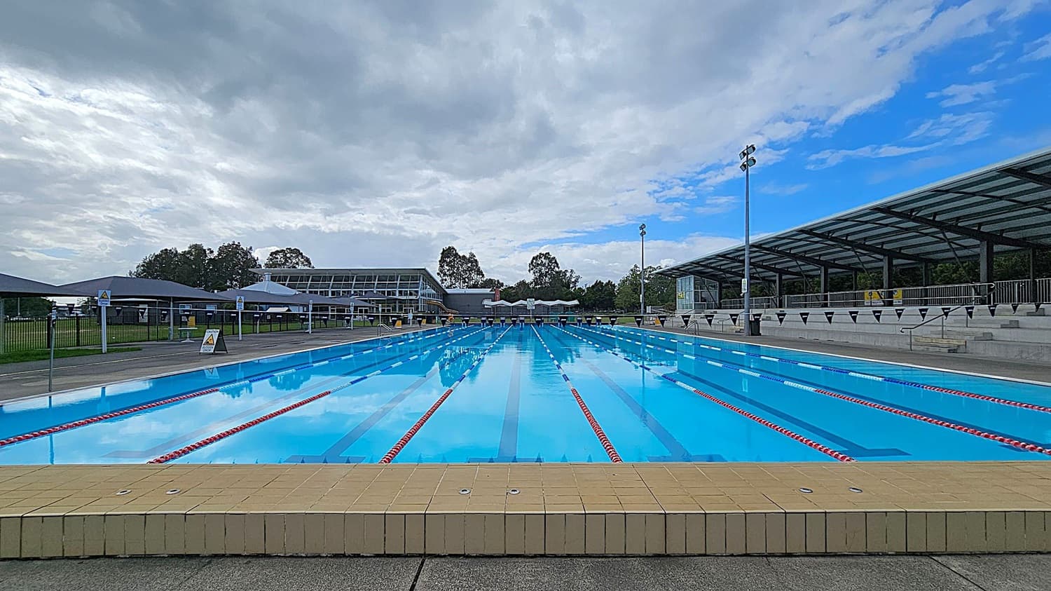 An outdoor swimming pool with clear blue water and red lane dividers stretches into the distance. On the right, there is a covered seating area with a metal railing and steps. On the left, a fence and several shaded structures are visible. In the background, there are trees and a large building with a glass facade. The pool deck is made of concrete, and the pool edge is lined with yellow tiles.