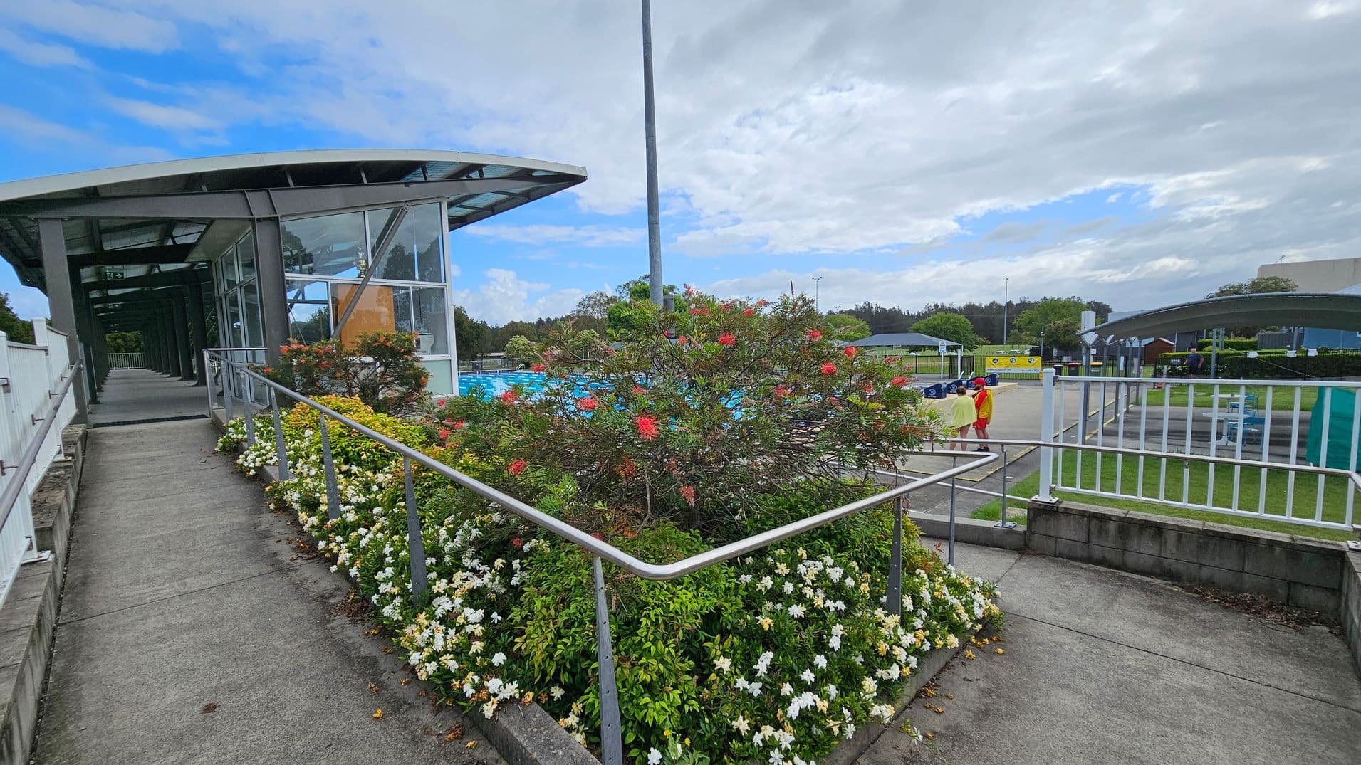 A walkway with a metal railing on the left leads to a glass building with a sloped roof. In the centre, a garden bed features a bush with red flowers and white blossoms at the base. On the right, two people in colourful clothing stand near a fenced area with a pool visible in the background. The ground is paved, and there are grassy areas with trees and additional structures in the distance.