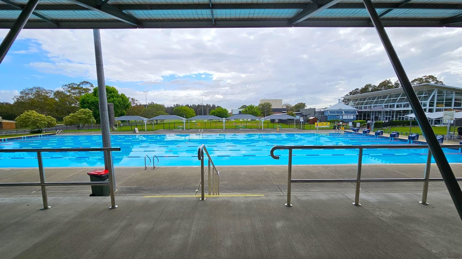 An outdoor swimming pool with clear blue water is in the centre, surrounded by a concrete deck. Metal railings and a bin are in the foreground. In the background, there are green trees and a grassy area. On the right, a modern building with large glass windows is visible. The ceiling is a metal structure with beams.