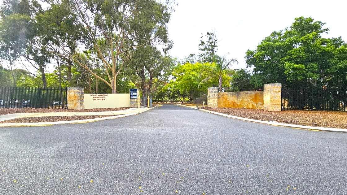 A road leading to the entrance. The gate is made of a huge brick structure. The left side features the facility name along with another banner. A pedestrian gate is also available on the left. A black steel fence borders the front. Tall trees surround the area.