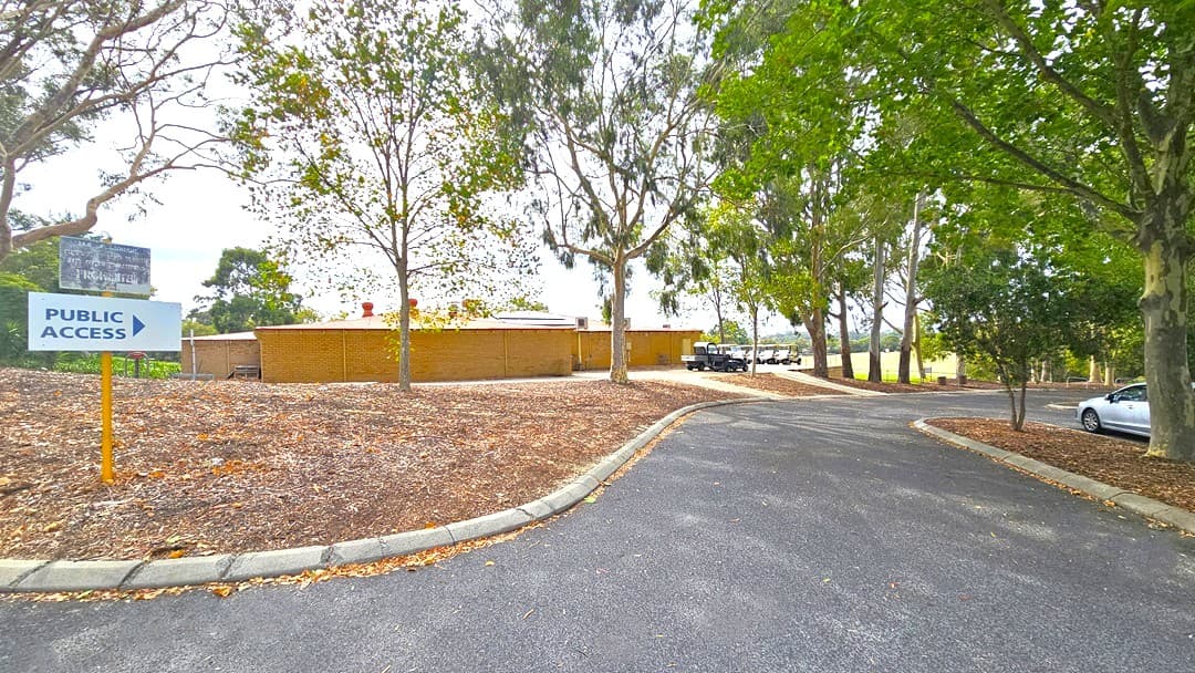 The road curves to the right, with a silver car visible near the curb. To the left, there is a sloping hill covered with dry leaves and tall trees. A sign reading "public access" is visible. In the middle of the road, two pathways elevate to the left. On the left, there is a low-rise building with a brown brick facade, and golf carts are parked in front of it.