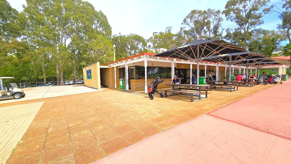 The facility building, constructed with brown bricks, stands on the right. In front, there is a veranda and a shaded area with picnic tables and chairs under large black umbrella shades. People are seated underneath. To the left, there is a spacious area with a cream brick wall bearing signage. A golf cart is parked nearby. The floor transitions from white concrete to brown tiles and red cement tiles. Tall trees surround the area.