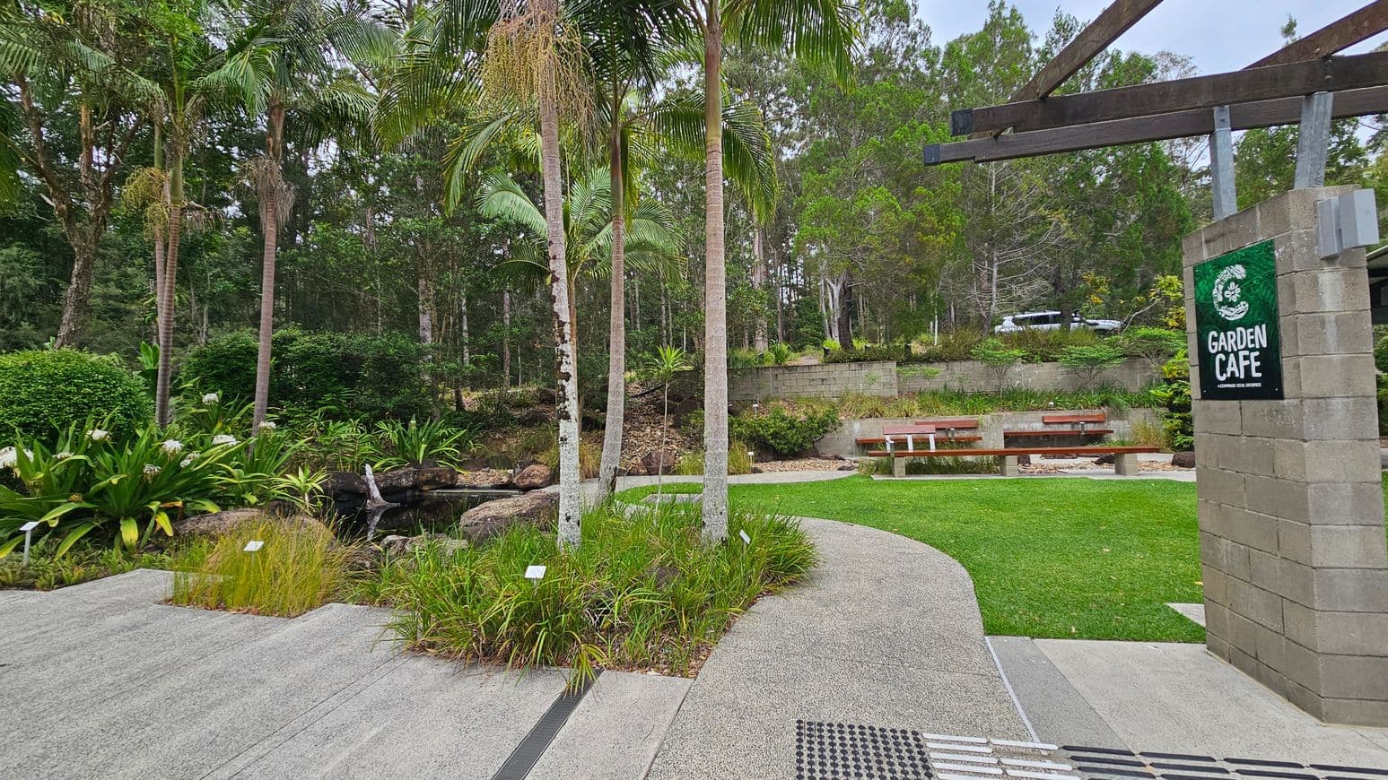 A garden scene with a modern building on the right, featuring a sloped roof and wooden columns. A winding path leads to the building, bordered by green grass and various plants. In the foreground, a concrete path intersects with the garden path. On the left, a black fence with a red "Tank Suction" sign and a fire hydrant is visible. A metal lamppost and a disabled parking sign are also present. Tall trees fill the background, creating a lush, wooded area.