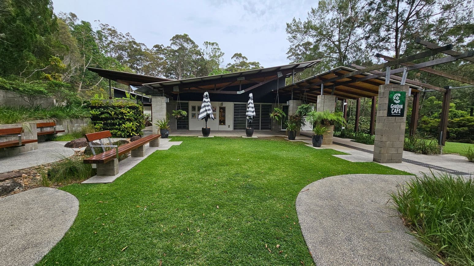 A garden café with a modern design, featuring a building with a slanted roof in the centre. The foreground has a well-maintained grassy area with a curved concrete path. On the left, there are wooden benches and a stone wall with lush greenery. On the right, a pergola with a "Garden Café" sign and potted plants. The background is filled with tall trees and dense foliage.