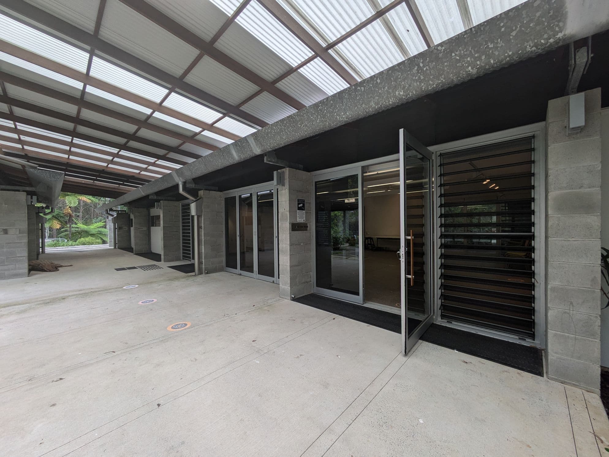 A covered walkway with a corrugated translucent roof. On the right, there are glass doors with a metal frame, one of which is open, and louvred windows beside them. The walls are made of grey concrete blocks. The floor is smooth concrete with circular markers. In the background, there are green plants and trees visible through the open space.
