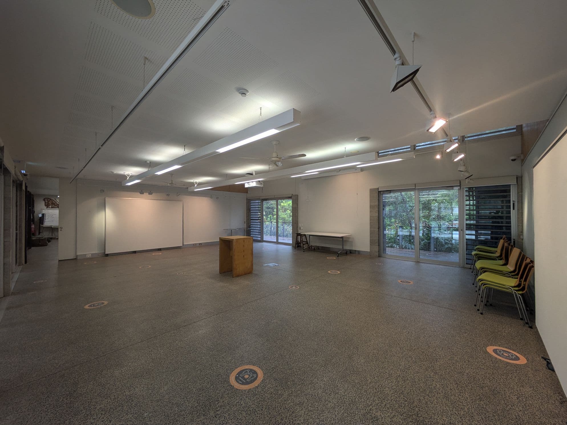 A spacious room with a speckled floor and a white ceiling featuring recessed lighting. On the left, there are large whiteboards and a small wooden podium in the centre. The right side has a stack of green chairs and large windows with a view of greenery outside. A table is positioned near the windows. Circular floor markers are evenly spaced across the room.