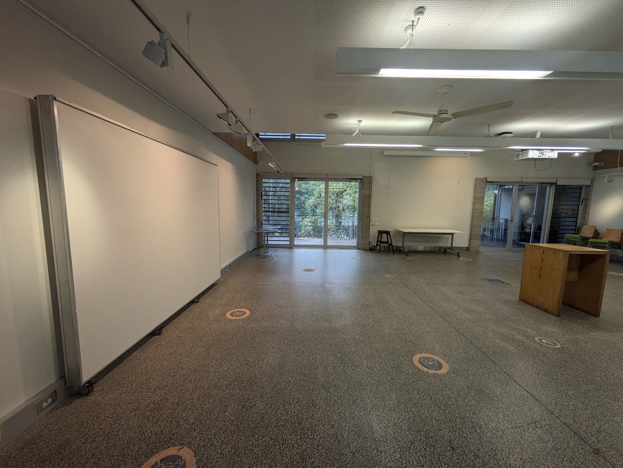 A spacious room with a speckled floor and a white ceiling featuring fluorescent lights and ceiling fans. On the left, there's a large whiteboard on wheels. The back wall has glass doors and windows, revealing greenery outside. In the centre, there's a table and a stool. On the right, a wooden podium and stacked chairs are visible. Circular markers are scattered on the floor.