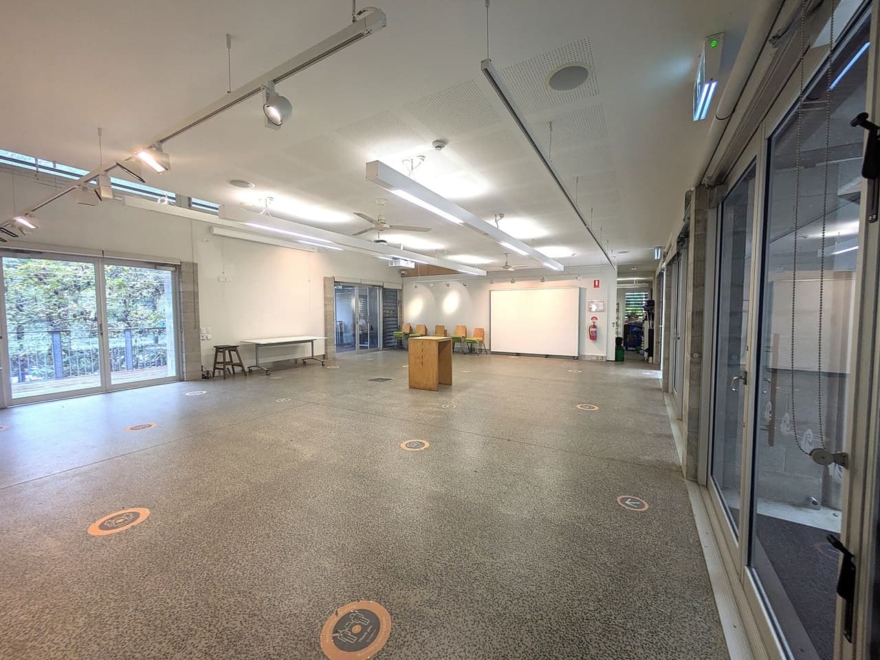 A spacious room with a speckled floor and a white ceiling featuring long fluorescent lights. On the left, large windows with a view of trees outside. A table and stool are against the left wall. In the centre, a wooden podium stands. The right side has glass doors leading to another area. At the back, a whiteboard and several chairs are visible, along with a fire extinguisher on the wall. Circular floor markers are scattered throughout the room.