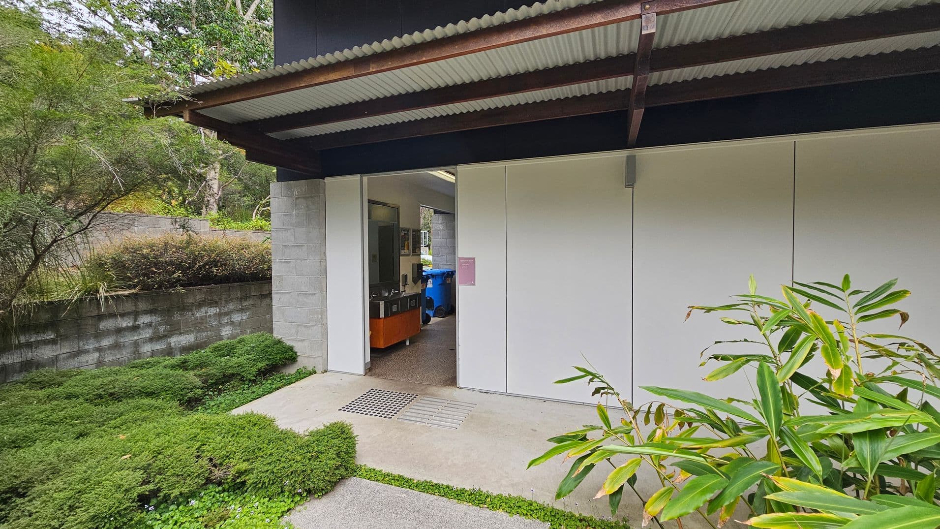 A building with a corrugated metal roof and white panelled walls. The entrance is open, revealing a room with a blue bin and a wooden cabinet inside. On the left, there's a grey brick wall with dense green shrubs and trees. On the right, there are tall green plants with broad leaves. The ground is a mix of concrete and grass.