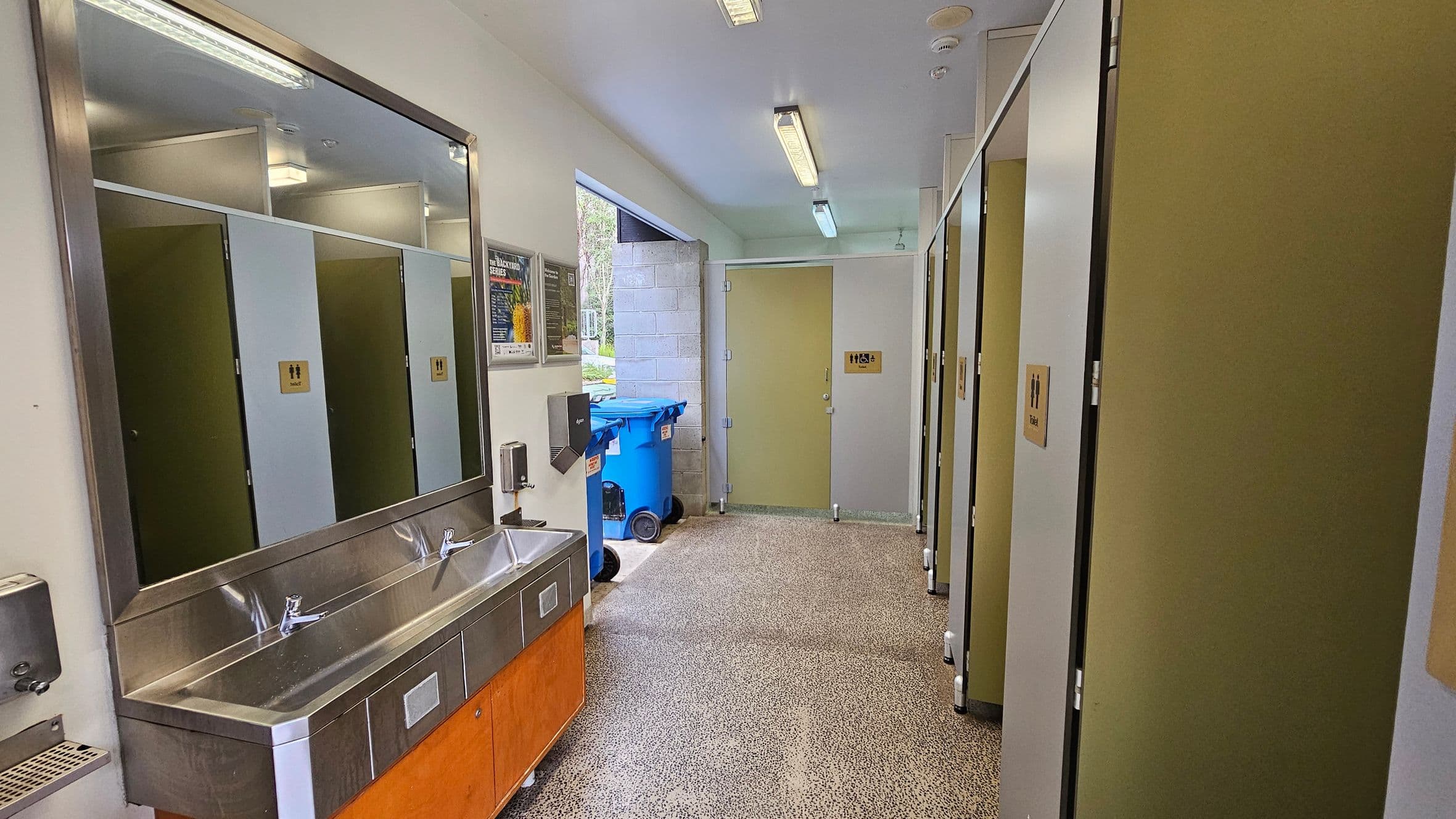 A public restroom with a long stainless steel sink and two taps on the left, beneath a large mirror. Above the sink, a paper towel dispenser and a soap dispenser are mounted on the wall. On the right, there are several toilet cubicles with green and white doors, each marked with a sign. The floor is speckled grey, and the ceiling has fluorescent lights. In the background, a partially open door reveals two blue recycling bins outside.