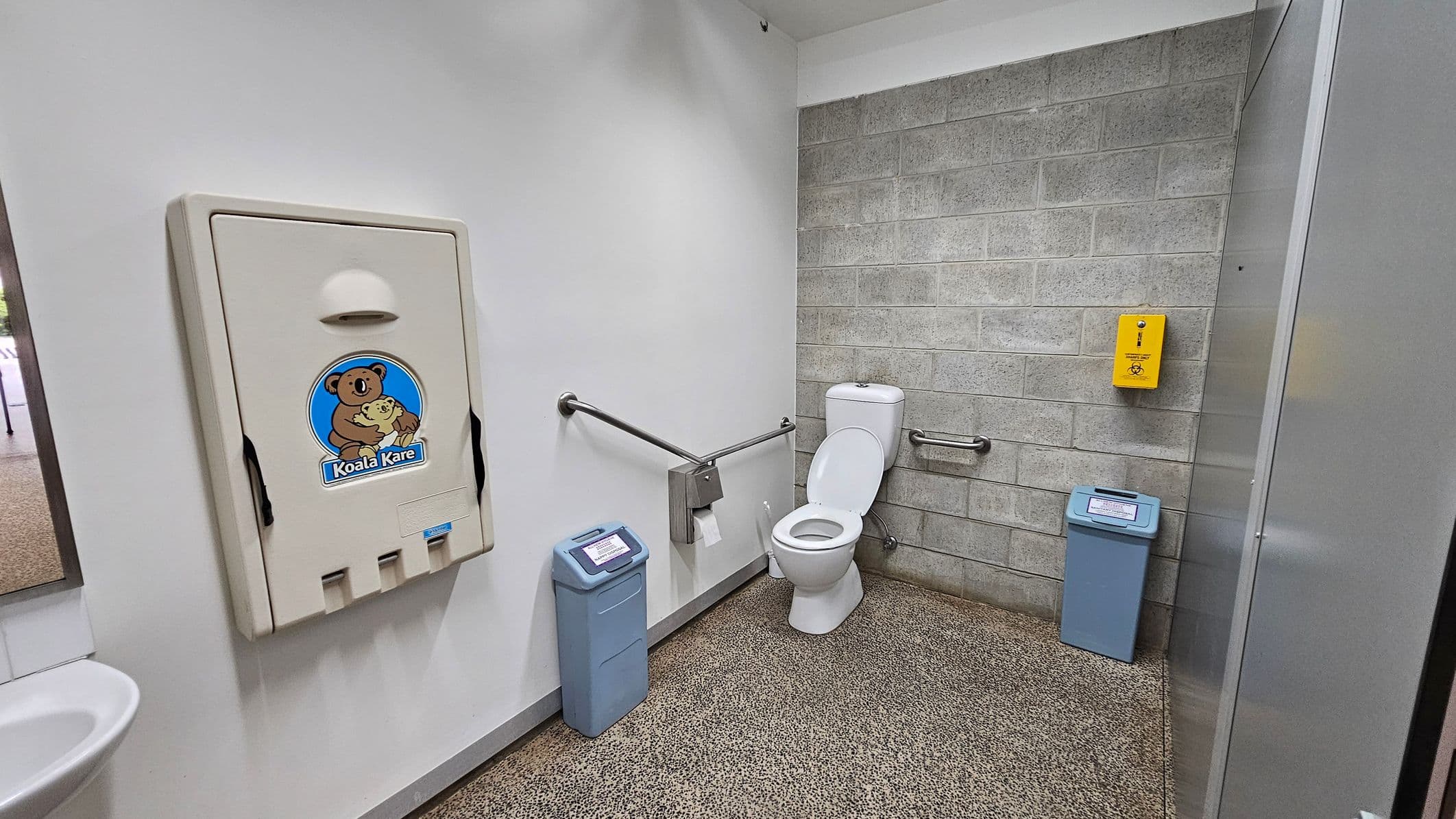 A public restroom with a baby changing station on the left wall, featuring a "Koala Kare" logo. The floor is speckled and the walls are a mix of white and grey brick. In the centre, there is a white toilet with grab bars on either side. A yellow sanitary disposal unit is mounted on the grey brick wall. Two blue bins are positioned near the toilet, one on each side. A small portion of a sink is visible on the far left.