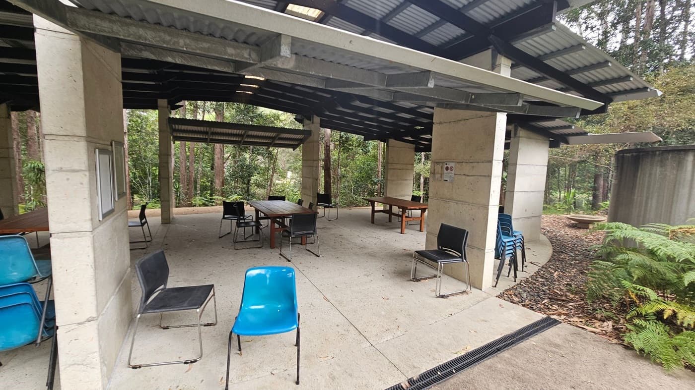 An outdoor covered area with a corrugated metal roof supported by concrete pillars. Several tables and chairs are scattered around, with some chairs stacked in blue and black. The floor is concrete, and the surrounding area is lush with trees and ferns. A large cylindrical water tank is visible on the right, and a small path covered in leaves leads into the greenery. The ceiling is open with visible beams.