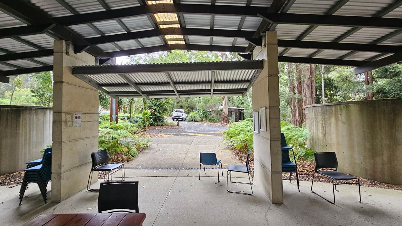 A covered outdoor area with a corrugated metal roof supported by concrete pillars. Several chairs are scattered around, with a stack of chairs on the left. The ground is concrete, and there are ferns and trees in the background. A white vehicle is parked on a paved path leading into a wooded area. A large cylindrical structure is partially visible on the right. The setting appears to be a natural, wooded environment.