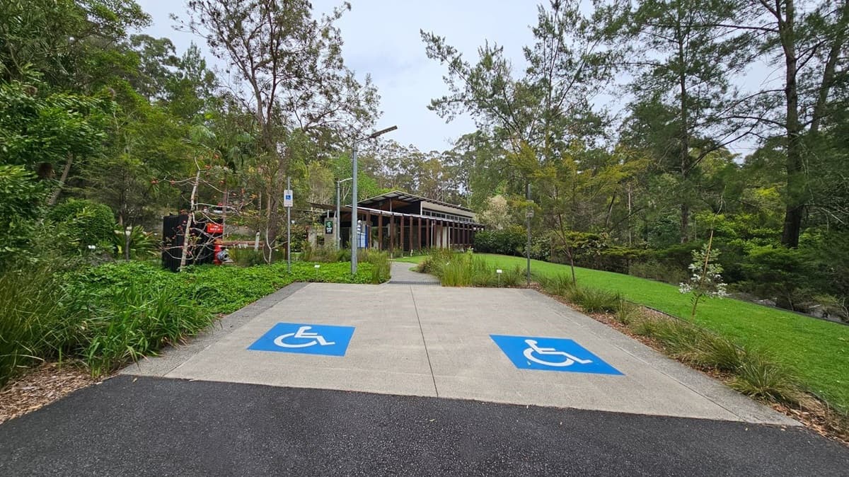 A pathway leads to a building with a slanted roof surrounded by trees and greenery. In the foreground, there are two parking spaces marked with blue disabled symbols. On the left, a red and black structure is partially visible among the foliage. The area is lush with grass and various plants, and a lamppost stands near the path.