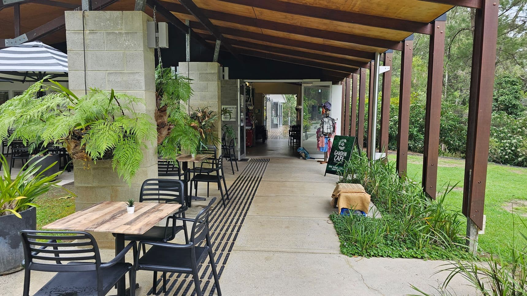 Outdoor café with wooden tables and black chairs on the left. Hanging ferns and potted plants decorate the area. A striped umbrella is partially visible. On the right, a walkway with a green lawn and a row of wooden beams. A sign reads "Garden Café" near the entrance, which is open with glass doors. Inside, more seating and a display with clothes and accessories. The ceiling is wooden with exposed beams.
