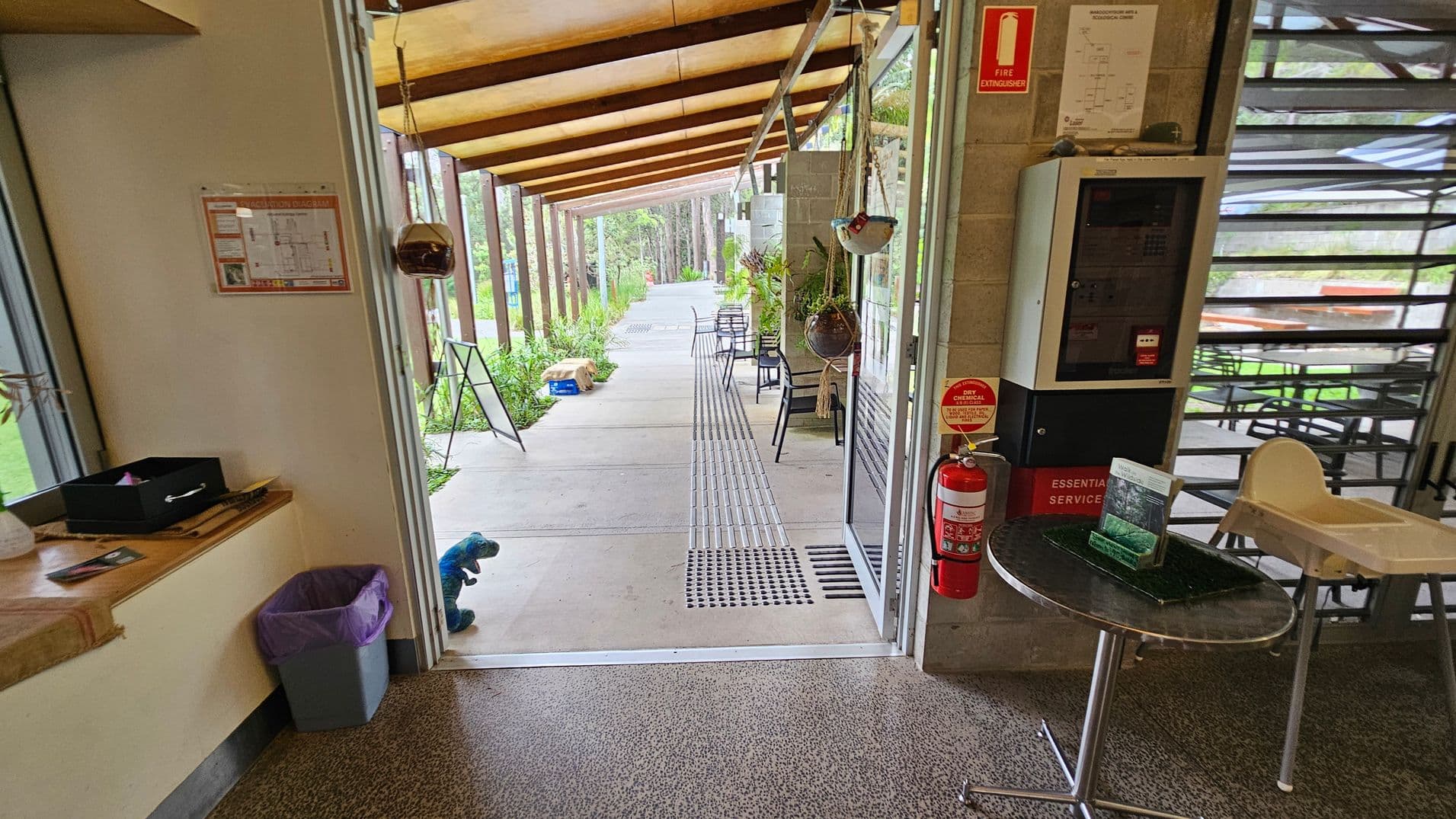 A view from inside a building looking out through an open door. On the left, there's a wall with a fire evacuation diagram and a small shelf with a black box and a bin below. On the right, a fire extinguisher is mounted on the wall next to a control panel. A small round table holds brochures, and a high chair is nearby. Outside, a covered walkway with hanging plants and chairs leads to a path surrounded by greenery. The floor is speckled, and the ceiling has wooden beams.