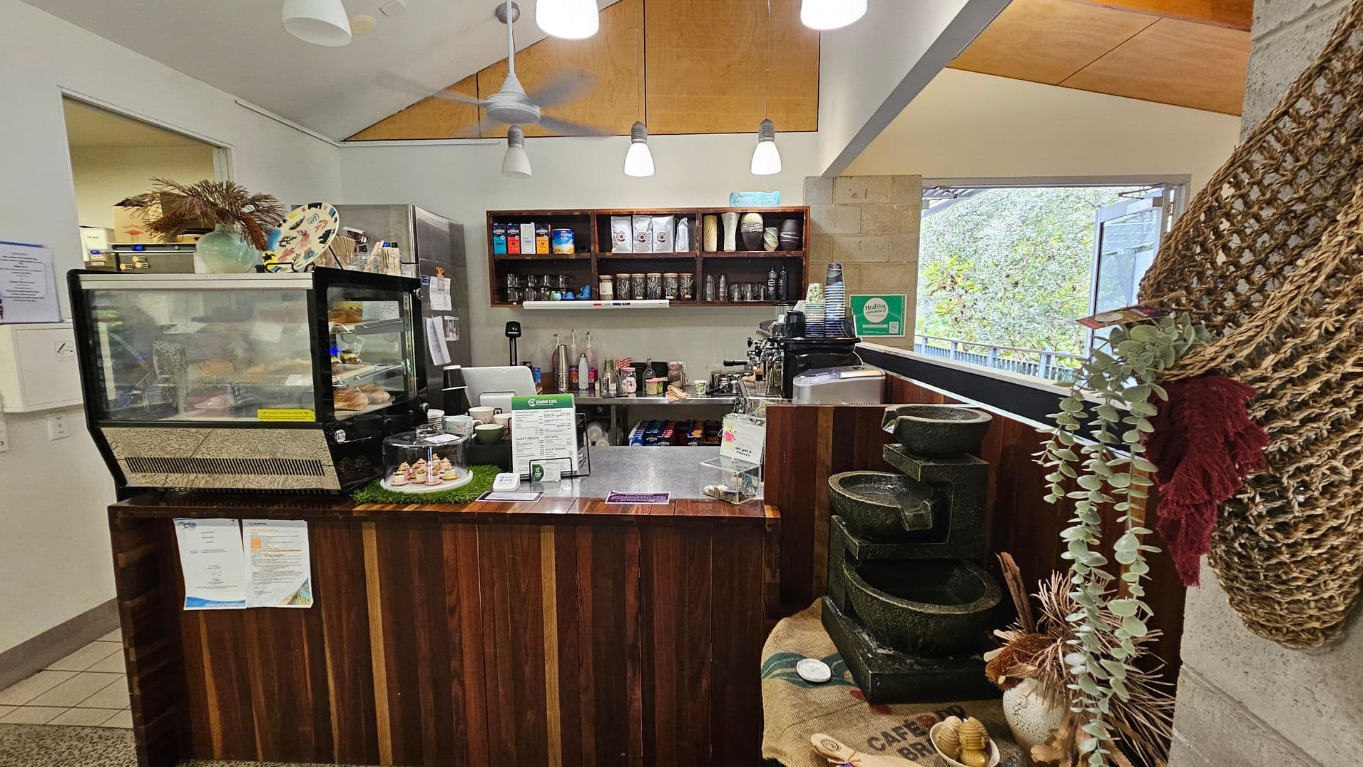 A small café counter with a wooden front. On the left, there's a glass display case with pastries and a decorative vase on top. In the centre, a menu and a card machine are on the counter. Behind, shelves hold coffee supplies and jars. On the right, a coffee machine and stacked cups are visible. A decorative net with greenery hangs on the far right. The ceiling has pendant lights, and a large window in the background shows greenery outside.