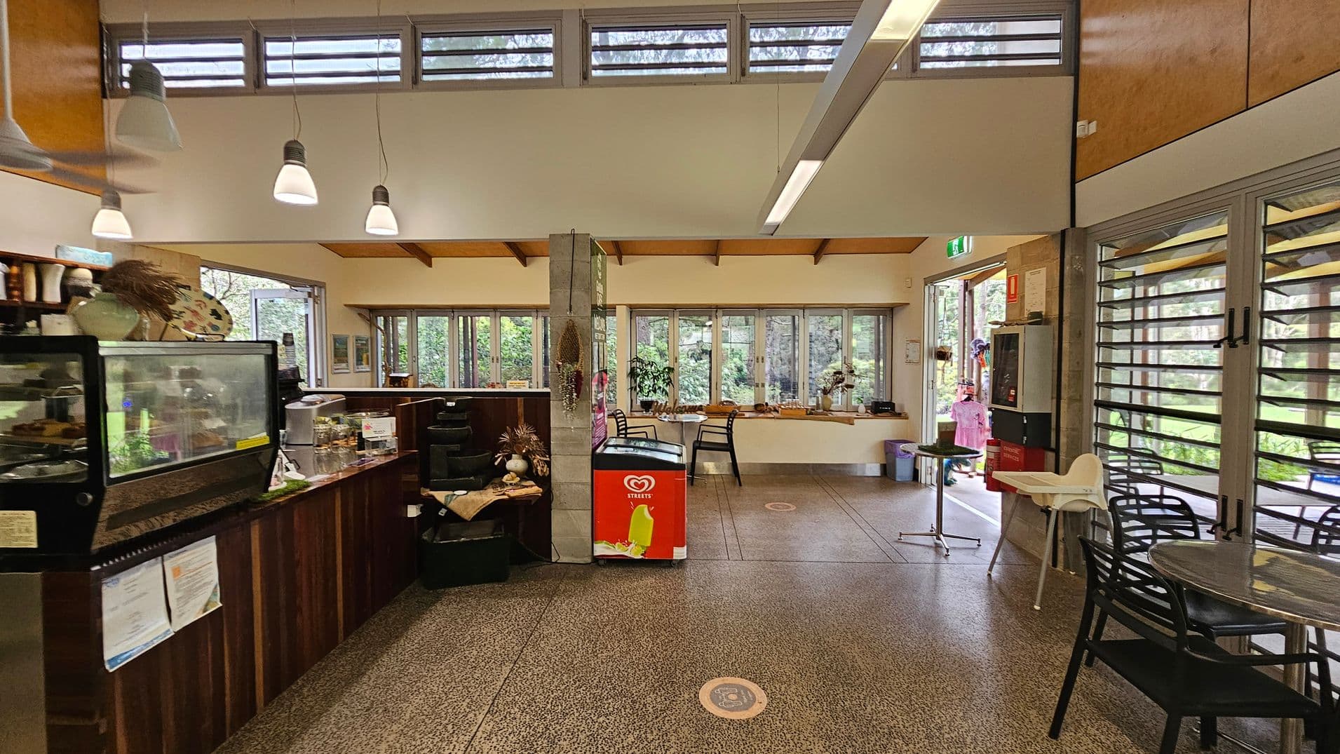 Interior of a café with a counter on the left featuring a glass display case and various items. In the centre, a red ice cream freezer is positioned against a column. The right side has tables and chairs, with large windows and glass doors providing a view of greenery outside. The ceiling is high with pendant lights, and the floor is speckled. The space is open and well-lit, with natural light streaming in through the windows.