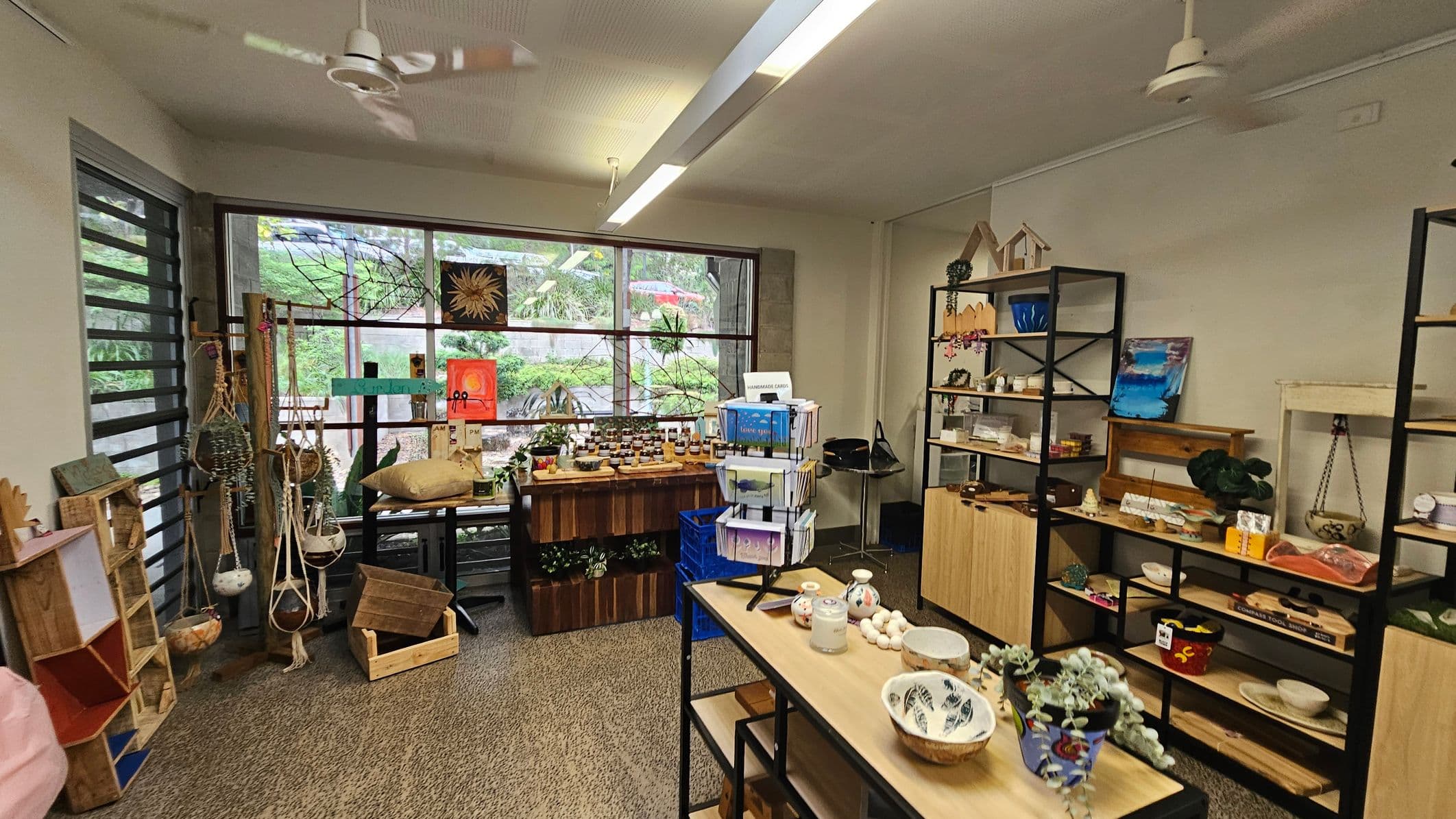 A small shop interior with various handmade items. On the left, there are hanging planters and wooden crates. The centre features a table with jars and a rotating card stand. The right side has shelves displaying pottery, artwork, and decorative items. Large windows in the background reveal greenery outside. The ceiling has two white fans, and the floor is carpeted.