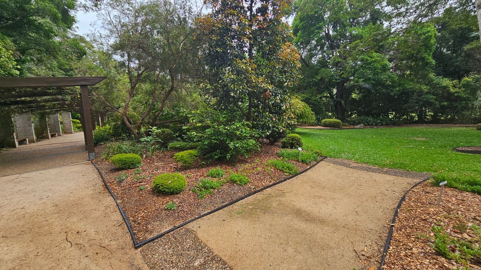 A garden scene with a sandy path curving through the centre. On the left, a wooden pergola with climbing plants leads to a shaded area. In the middle, a variety of shrubs and small trees are planted in a mulched bed. On the right, a well-maintained lawn is bordered by more trees and bushes. The background is filled with dense greenery, creating a lush, natural setting.