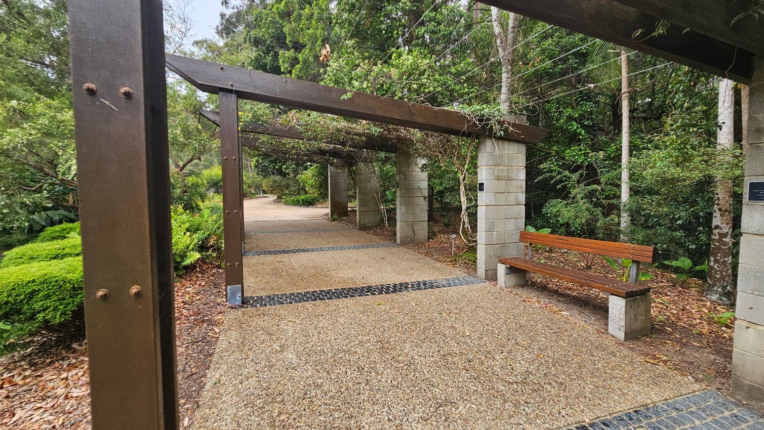 A gravel path leads through a garden with wooden pergolas supported by concrete pillars. On the right, a wooden bench is positioned next to a pillar. Lush greenery, including ferns and trees, surrounds the path. The pergolas are covered with climbing plants, and the ground is scattered with fallen leaves.