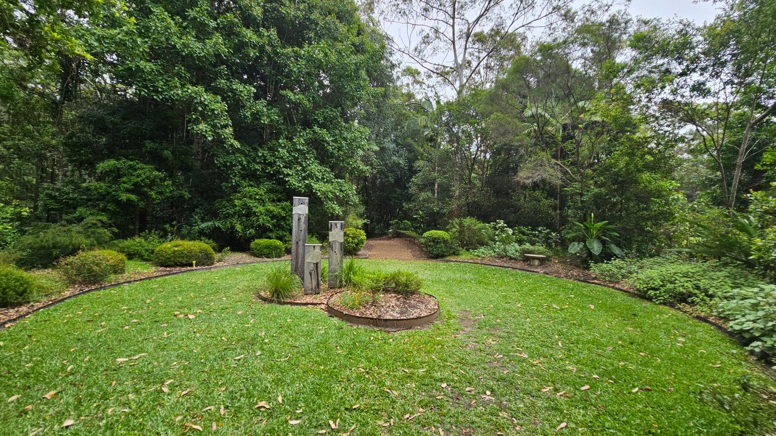 A lush garden with a circular grassy area in the foreground. In the centre, there are three wooden posts surrounded by small plants. The background is dense with various trees and shrubs, creating a natural border. A narrow dirt path leads into the wooded area, flanked by more greenery. A small stone bench is visible to the right, partially obscured by foliage. The scene is vibrant with shades of green.