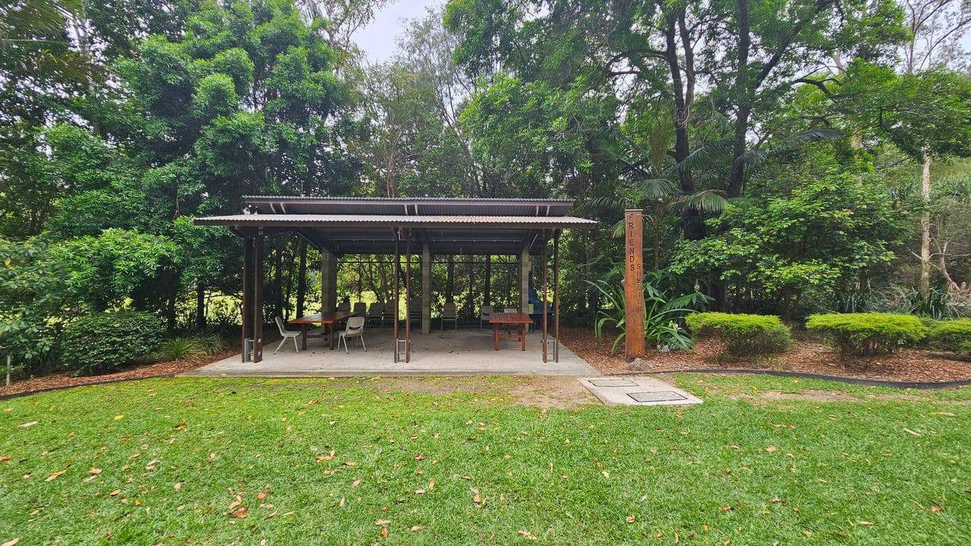 A small open shelter with a corrugated metal roof is situated in a lush, green park. Inside, there are several chairs and a table. To the right, a wooden post with the words "FRIENDS SHELTER" stands amidst dense foliage. The foreground features a well-maintained grassy area with scattered leaves. The background is filled with tall trees and bushes, creating a natural, serene setting.