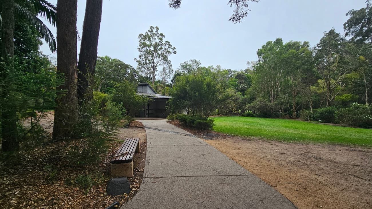 A paved path leads through a park with a wooden bench on the left, surrounded by tall trees and dense foliage. In the centre, a small building with a sloped roof is partially obscured by bushes. To the right, a well-maintained grassy area extends towards more trees in the background. The ground is a mix of pavement and dirt, with scattered leaves.