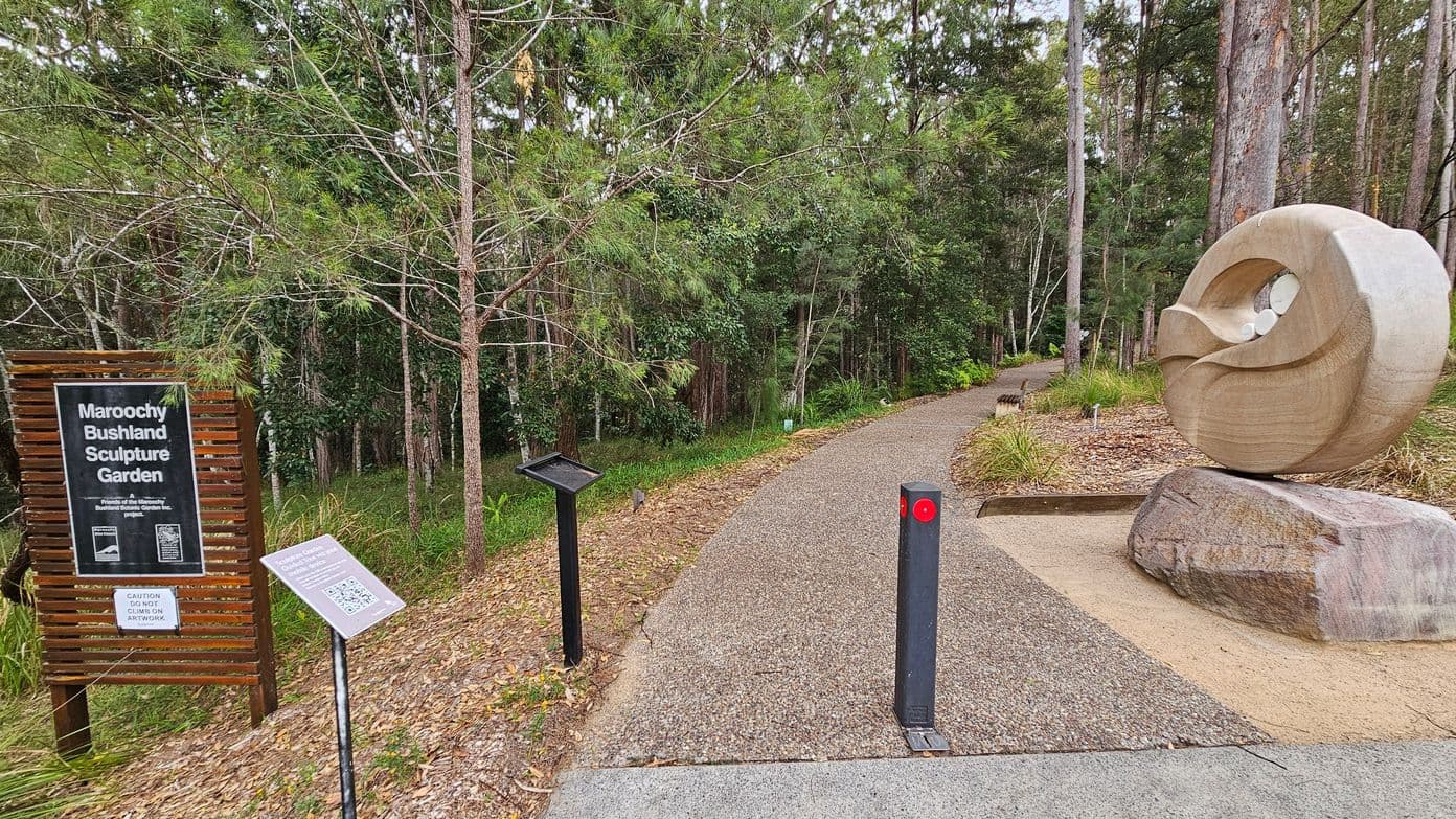 A pathway leads into a dense forest area on the left, with a sign reading "Maroochy Bushland Sculpture Garden" near the entrance. A large, abstract stone sculpture is positioned beside the path. On the right, there is a paved parking area with marked spaces. The ground is covered with mulch and the area is surrounded by tall trees and green foliage.