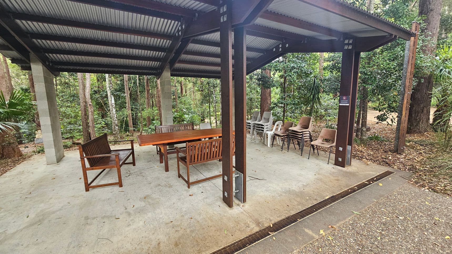 A covered outdoor area with a corrugated metal roof supported by wooden and concrete pillars. On the left, there are wooden benches and a table. On the right, several stacks of plastic and metal chairs are arranged. The ground is a concrete surface with some leaves scattered around. In the background, there is dense greenery with tall trees and bushes.