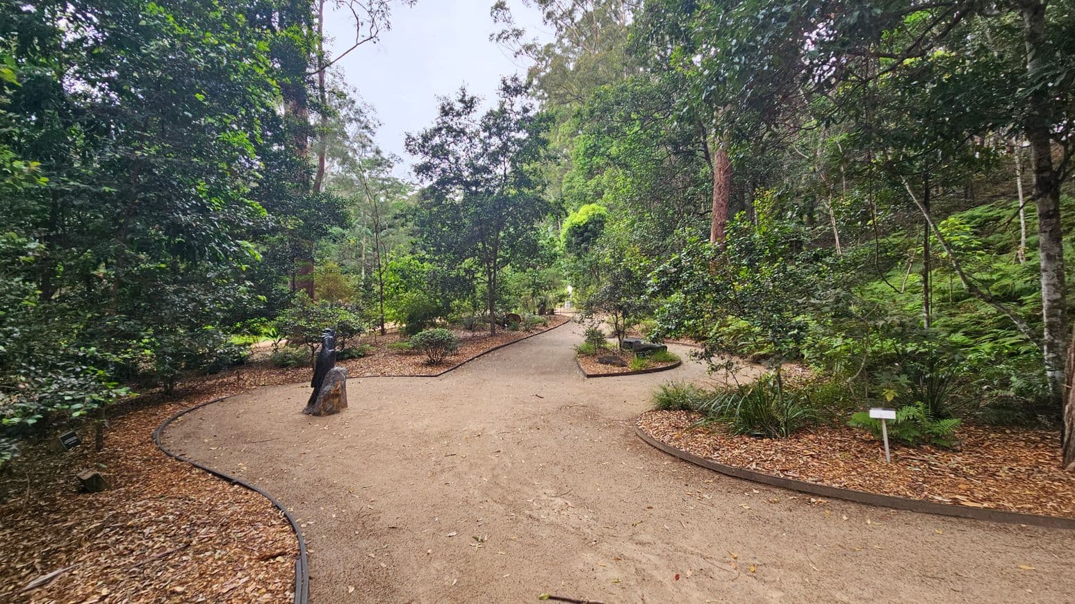 A winding dirt path in a lush garden setting, surrounded by dense greenery and tall trees. The path splits into two directions, with a small statue on the left side. The ground is covered with fallen leaves, and there are small shrubs and plants along the path edges. A sign is visible on the right, partially obscured by foliage. The background is filled with more trees and greenery, creating a serene, natural atmosphere.