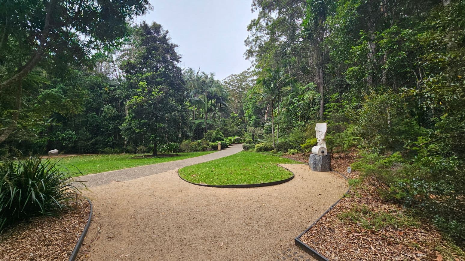 A winding path in a lush garden setting. On the left, dense green foliage and a grassy area. On the right, a modern sculpture on a stone pedestal surrounded by bushes. The path is bordered by a mix of grass and mulch. Tall trees and palms are visible in the background, creating a serene, natural atmosphere.