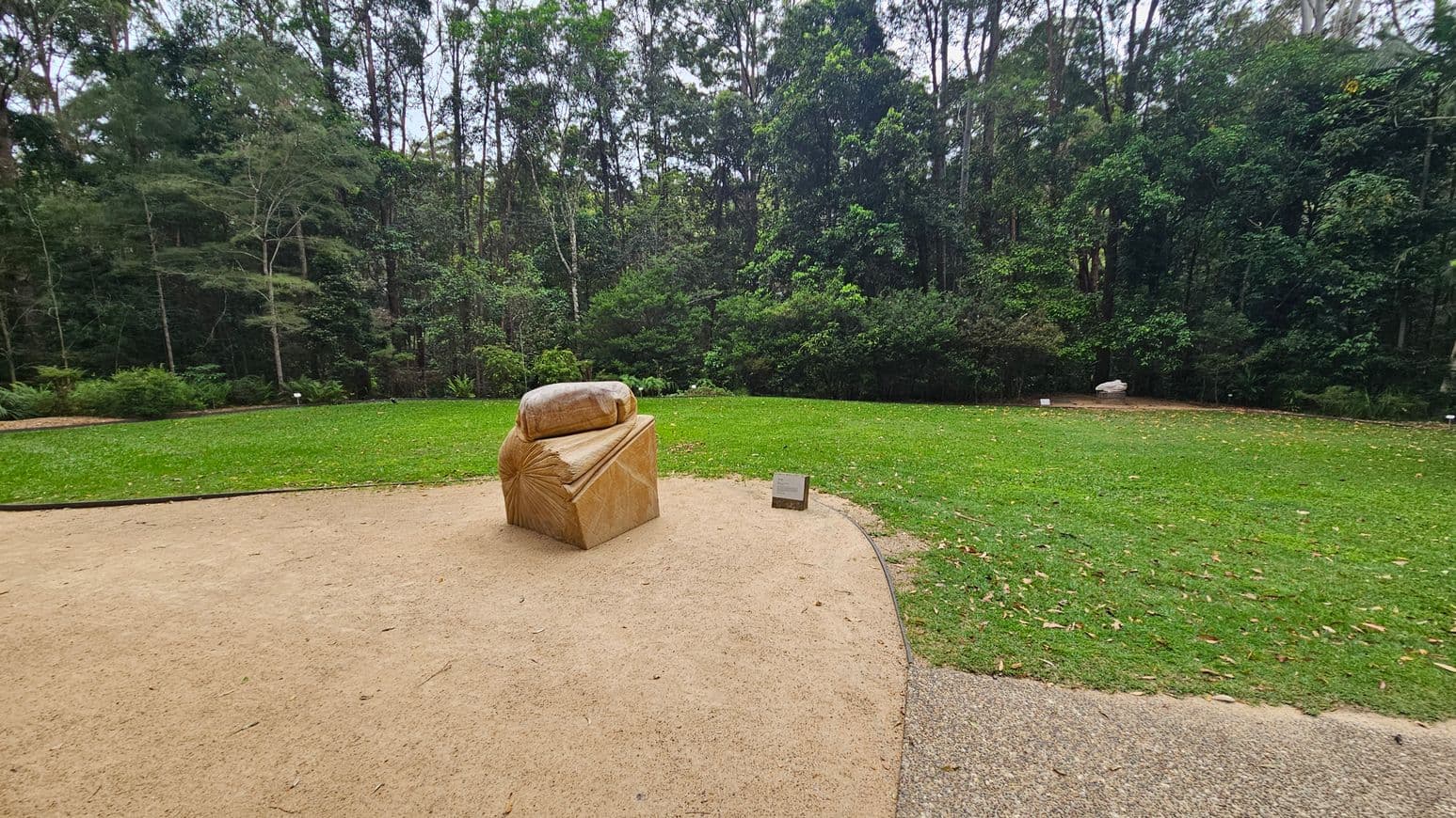 A stone sculpture resembling a draped cloth is positioned on a sandy area in the foreground. To the right, a small plaque is placed on the ground. The background features a lush green lawn bordered by dense trees with varying shades of green foliage. The scene is set in a natural, outdoor environment.