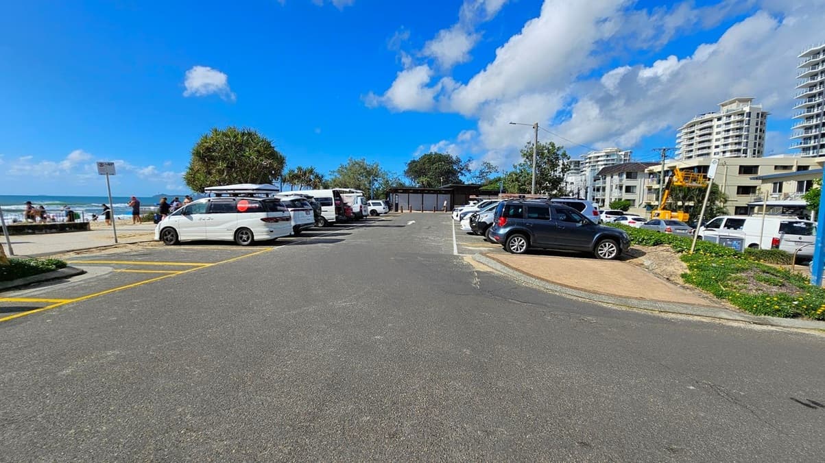A car park with several vehicles, including white vans and cars, is in the foreground. On the left, people are gathered near the beach, with the ocean visible beyond. In the background, there are trees and a small building. On the right, modern multi-storey buildings and a construction vehicle are visible. The ground is paved, and there are yellow parking lines.