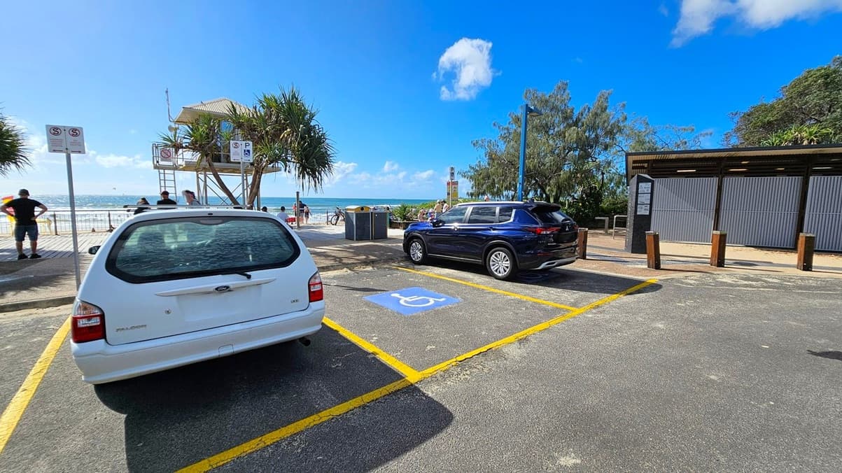 A coastal parking area with two cars parked in designated spaces. On the left, a white car is parked next to a sign indicating parking restrictions. In the centre, a blue car is parked in a space marked with a disability symbol. In the background, a lifeguard tower overlooks the beach, with people nearby. To the right, a building with closed shutters is visible. The ocean and trees are in the distance, and the ground is paved with visible yellow lines.
